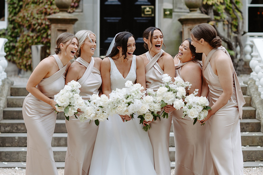 Bride and her five bridesmaids laugh together while holding white rose bouquets